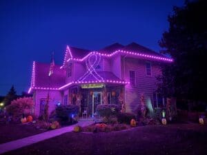 Brightly lit house in Fond du Lac with vibrant purple neon lights outlining the roof and a large ribbon design, surrounded by fall pumpkins and corn stalks