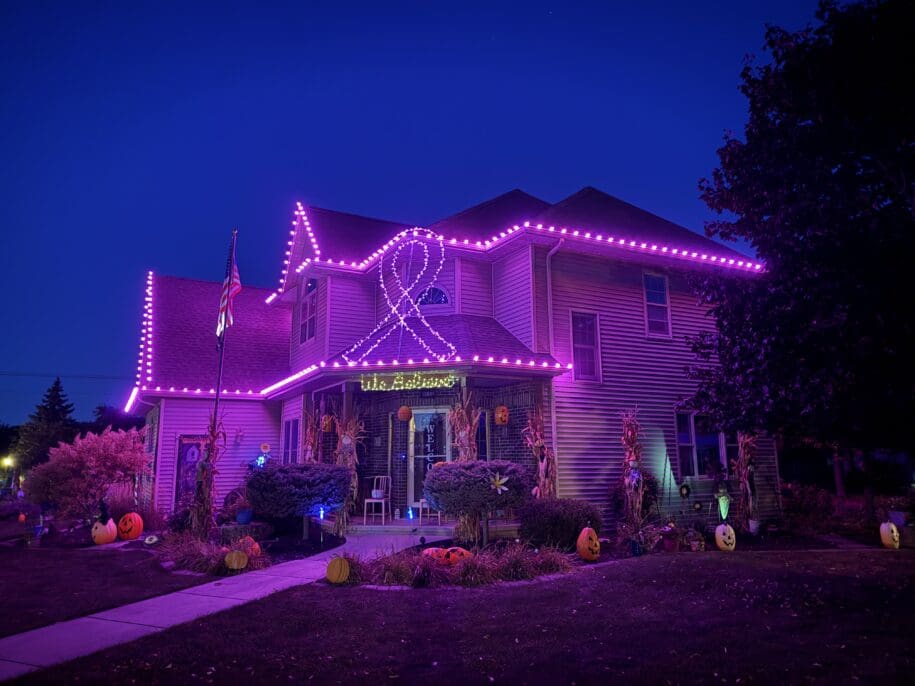 Brightly lit house in Fond du Lac with vibrant purple neon lights outlining the roof and a large ribbon design, surrounded by fall pumpkins and corn stalks