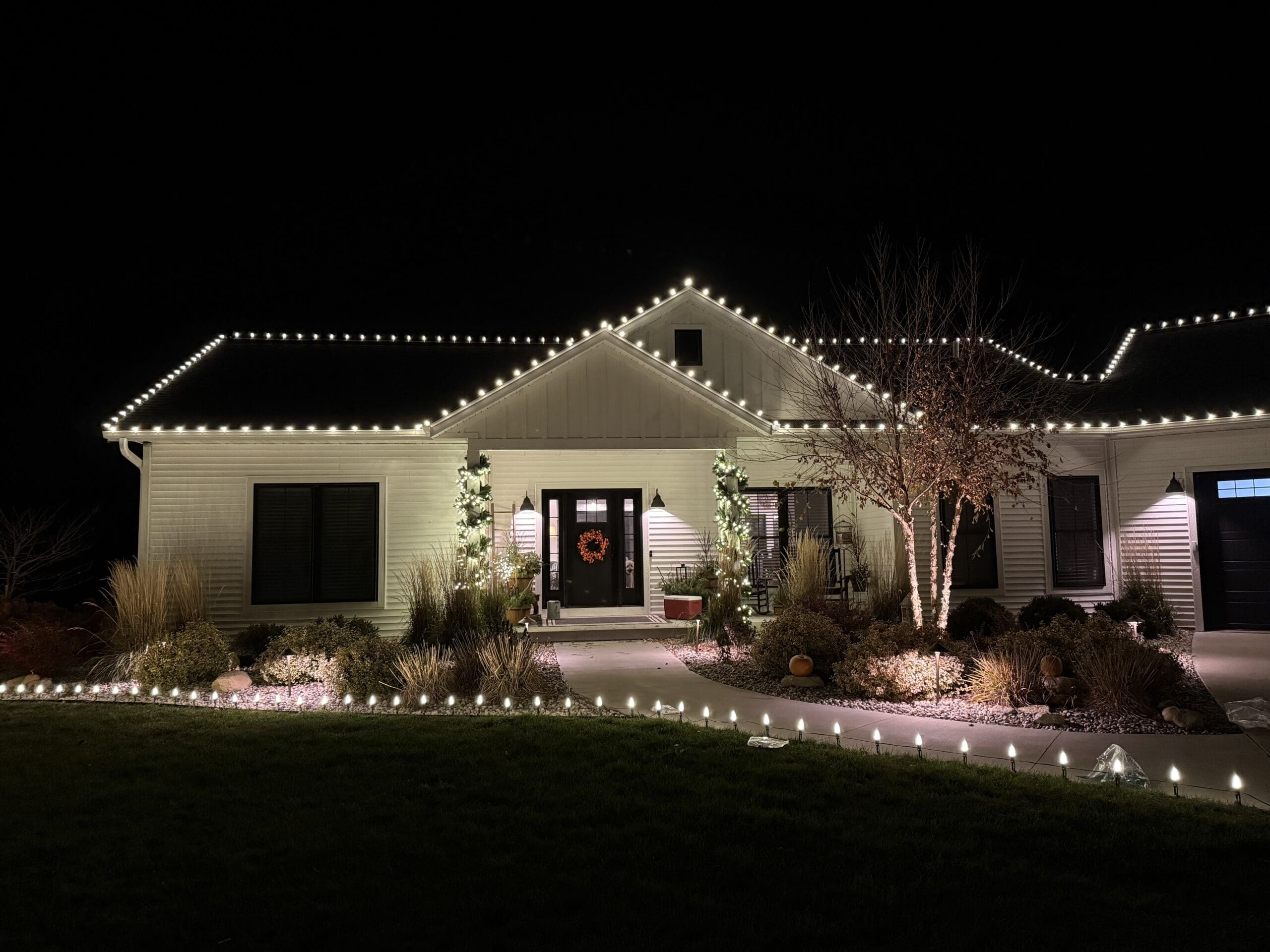 White single-story house with elegant holiday lighting along roofline and landscaping, featuring wreath on front door and illuminated shrubs at night