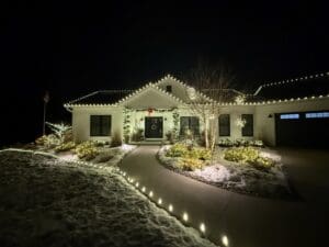 White ranch house with festive holiday lighting on roofline, decorated garland on dark front door, illuminated landscaping along curved driveway with snow in Fond du Lac Wisconsin