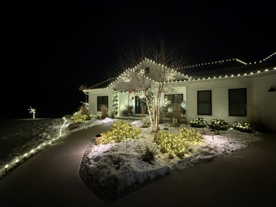 White residential home illuminated at night with holiday decorations, snow-covered landscaping, and glowing shrubs in Fond du Lac, Wisconsin